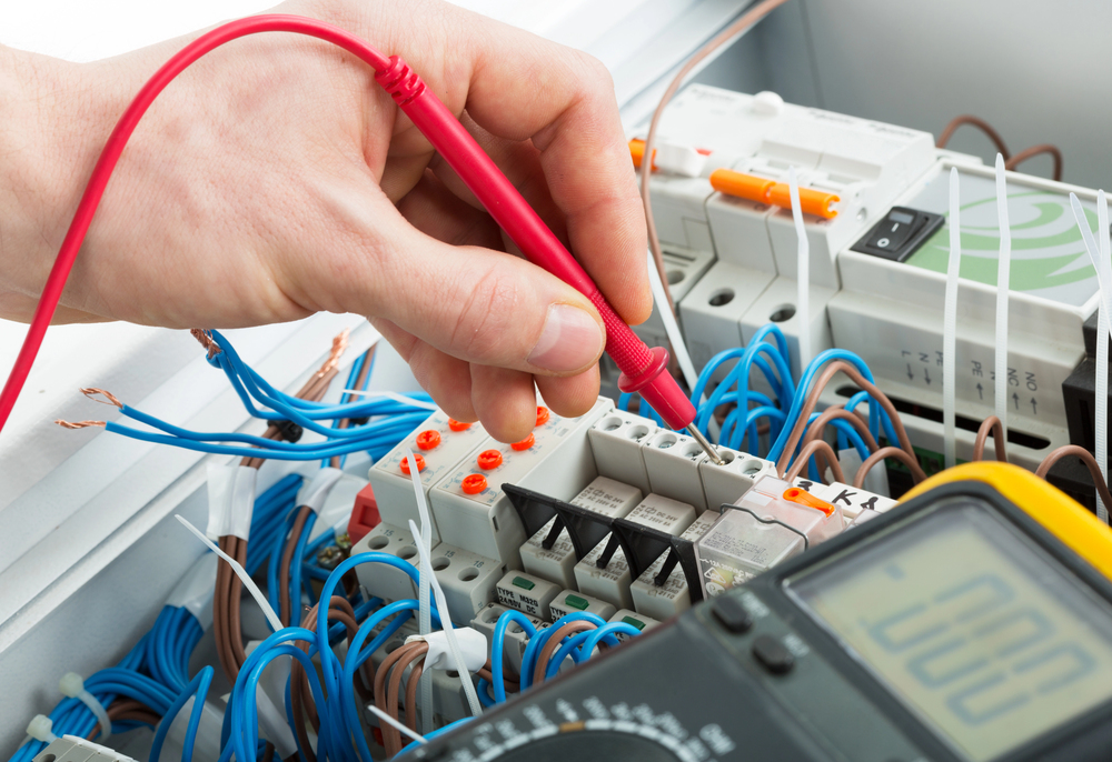 A hand uses a red multimeter probe to test electrical connections in a control panel with numerous blue wires and circuit components.