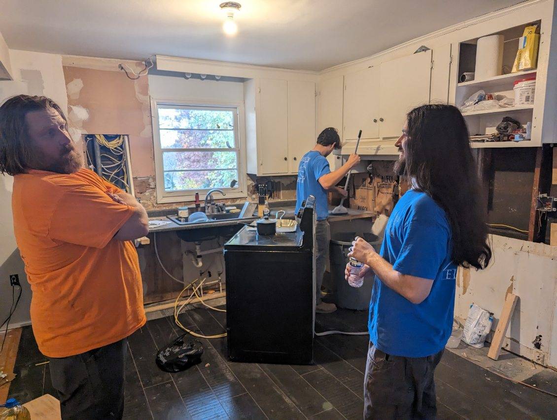 Three men work on a kitchen renovation; two stand and talk while one fixes plumbing under the sink. Cabinets and walls are partially dismantled.