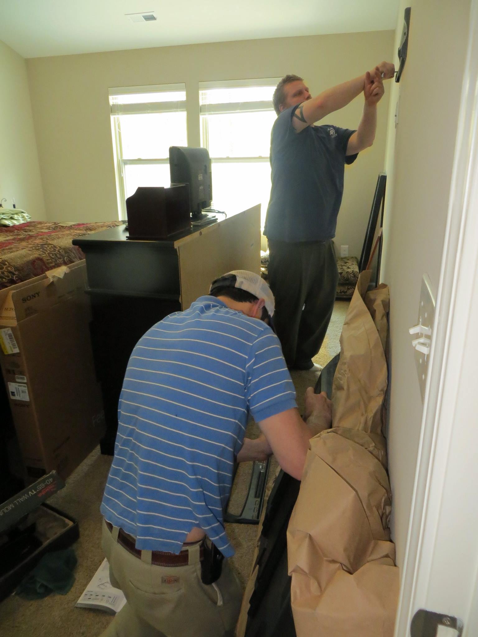 Two people work in a bedroom; one adjusts electrical wiring on the wall while the other kneels on the floor, working on a project near boxes and packing material.