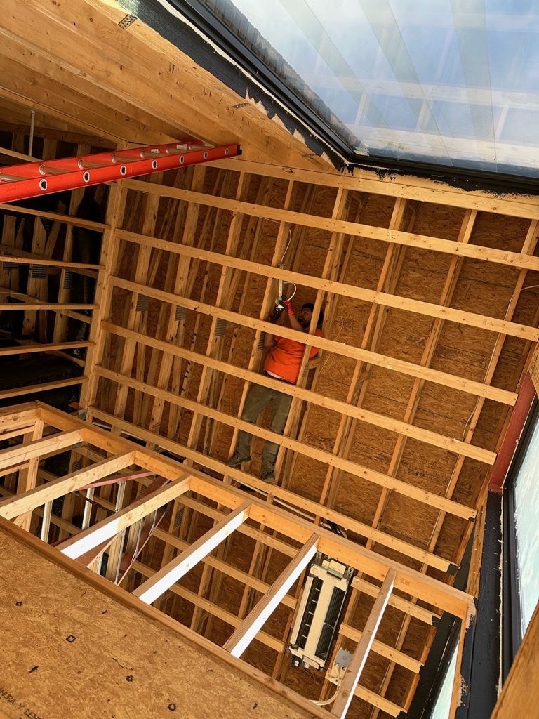 A construction worker in an orange shirt stands on a wooden frame, working on the ceiling structure inside a building under construction.