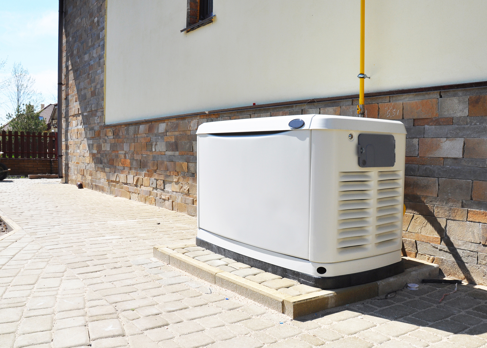 A standby generator installed on a concrete pad next to a house with stone siding, connected to a yellow utility pipe.