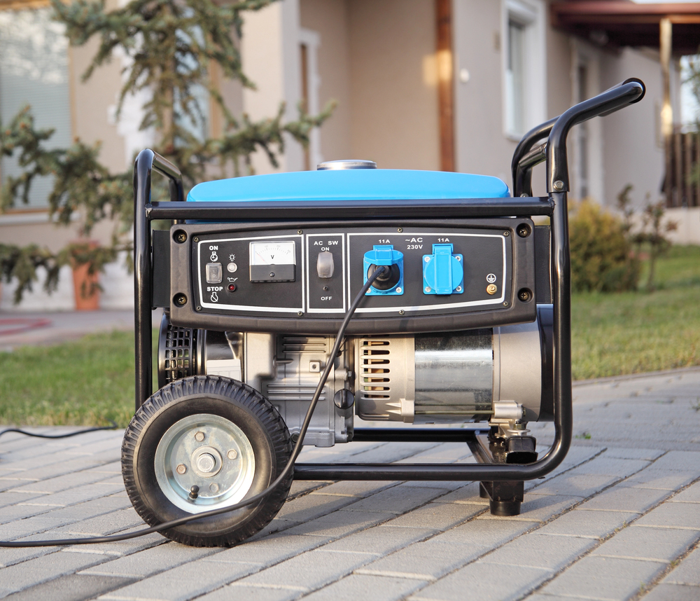 A portable gasoline generator with a blue cover is positioned on a paved walkway outside a house, connected with a power cord.