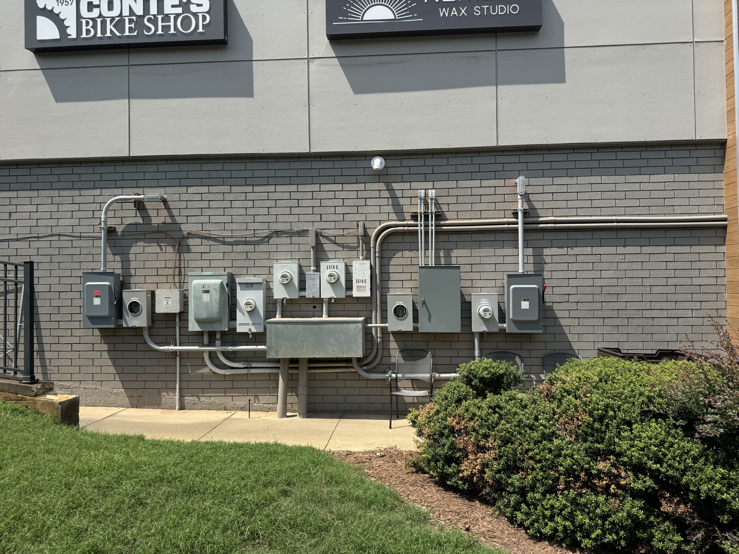 A row of electrical utility meters and breaker boxes mounted on a brick wall outside a building, with conduit pipes and a utility sink below. Shrubs and grass are in the foreground.