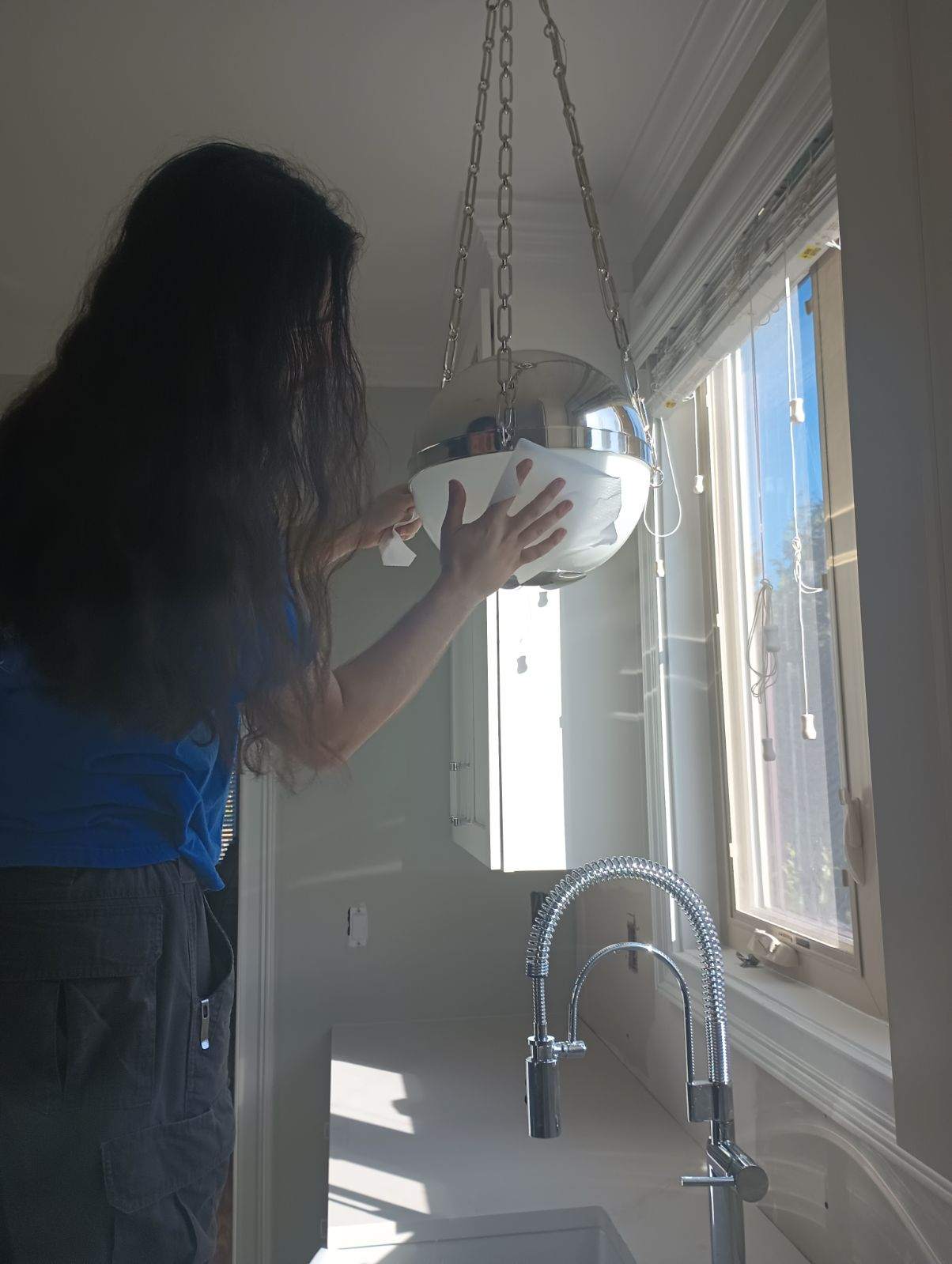 Person with long hair adjusts or installs a hanging light fixture above a kitchen sink near a window with sunlight streaming in.