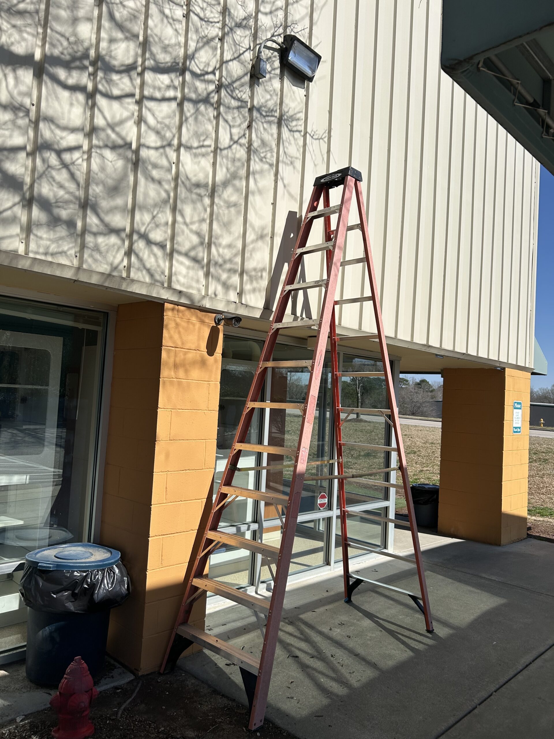 A red step ladder is positioned on a sidewalk, leaning against a yellow and beige building under a mounted light fixture.