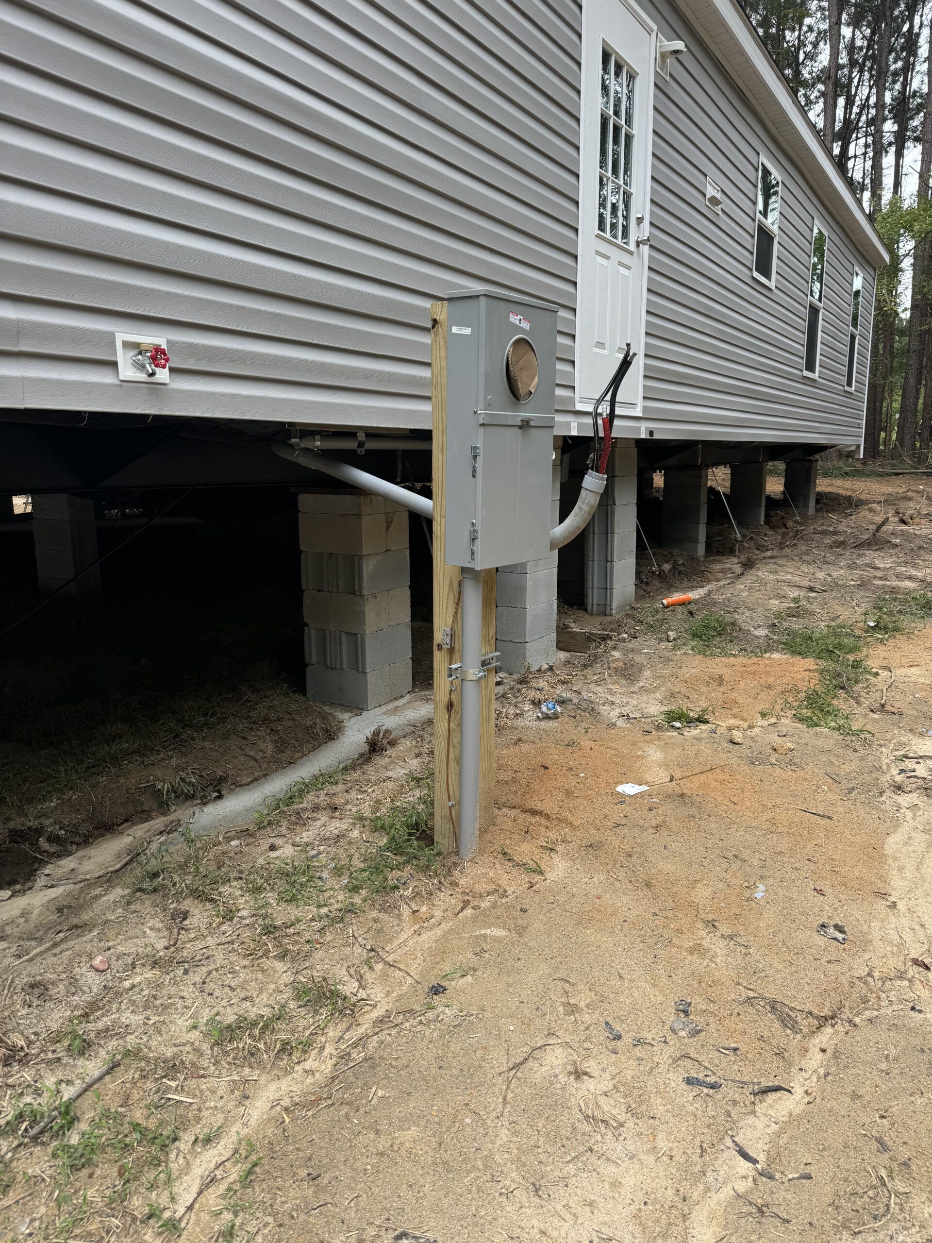 A raised manufactured home sits on concrete blocks with an electric meter base and service panel mounted on a wooden post in the foreground.