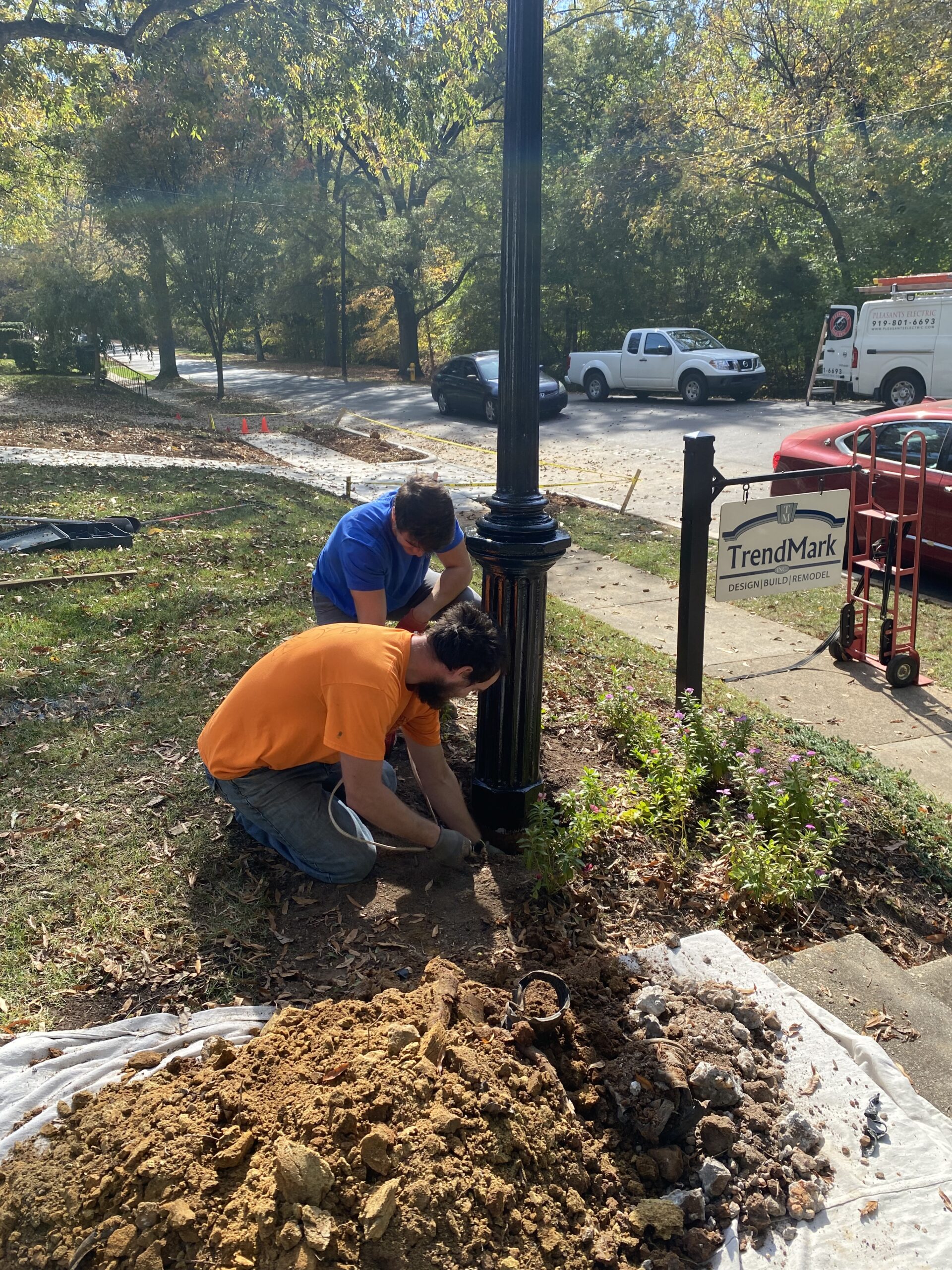 Two workers are installing or repairing a black lamppost beside a sidewalk, with a pile of dirt and tools nearby. A TrendMark sign is visible in the background.